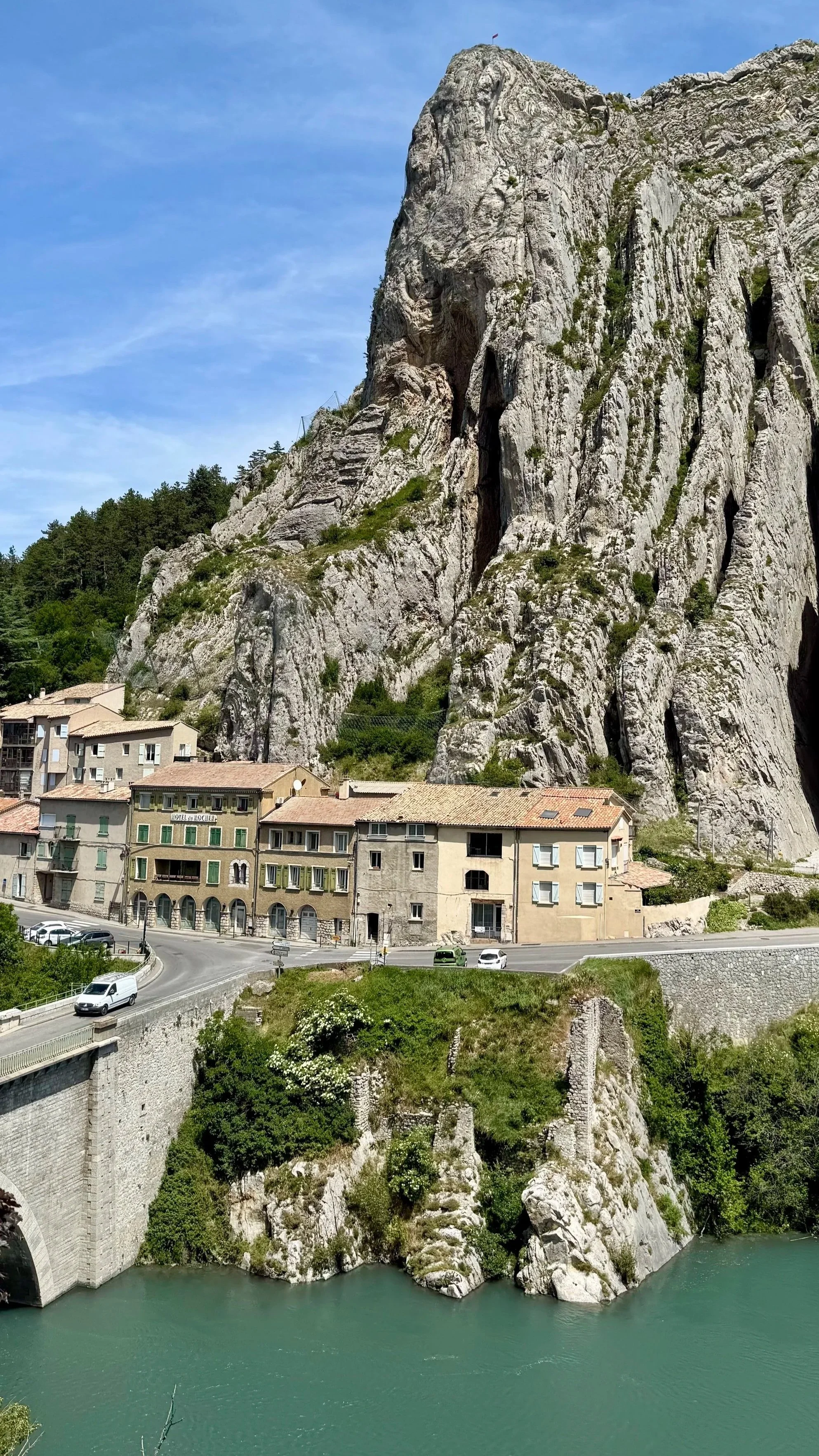 A scenic view of a large rocky mountain with buildings at its base, a curved road, and a river at the bottom with clear green water under a blue sky.