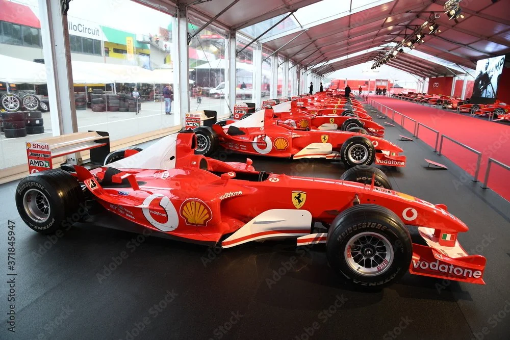 Multiple red Ferrari Formula 1 race cars displayed in a glass-walled exhibition tent with logos of sponsors, including Shell, Vodafone, and Bridgestone.