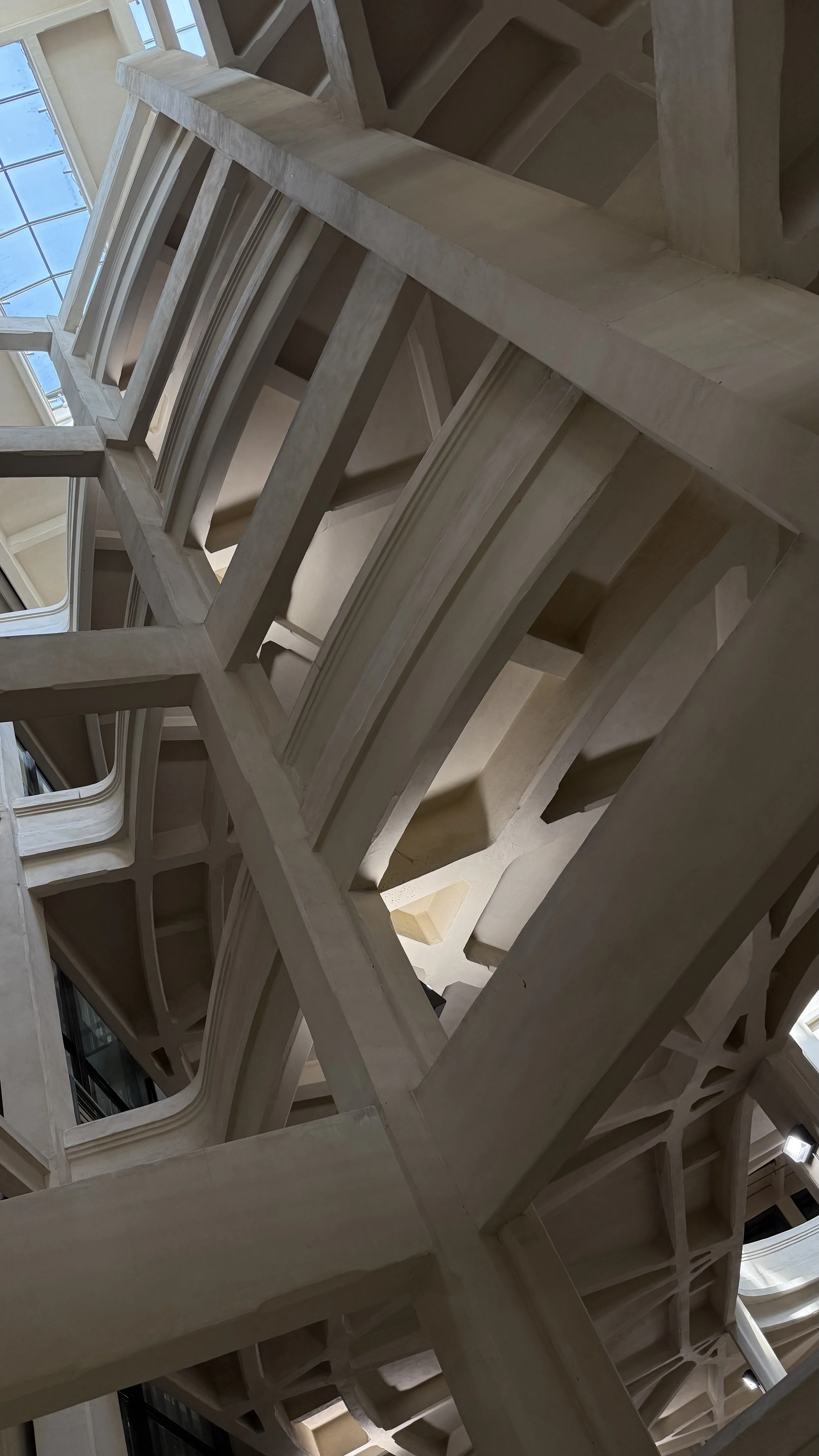 Interior view of a modern architectural staircase with multiple levels, featuring open spaces, beige-colored stone or concrete structures, and glass windows at the top letting in natural light.