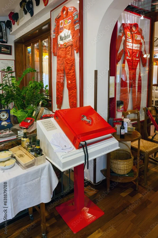 Display case with a racing suit and memorabilia, a table with bottles, food, and a book with the Ferrari logo, inside a restaurant or café.