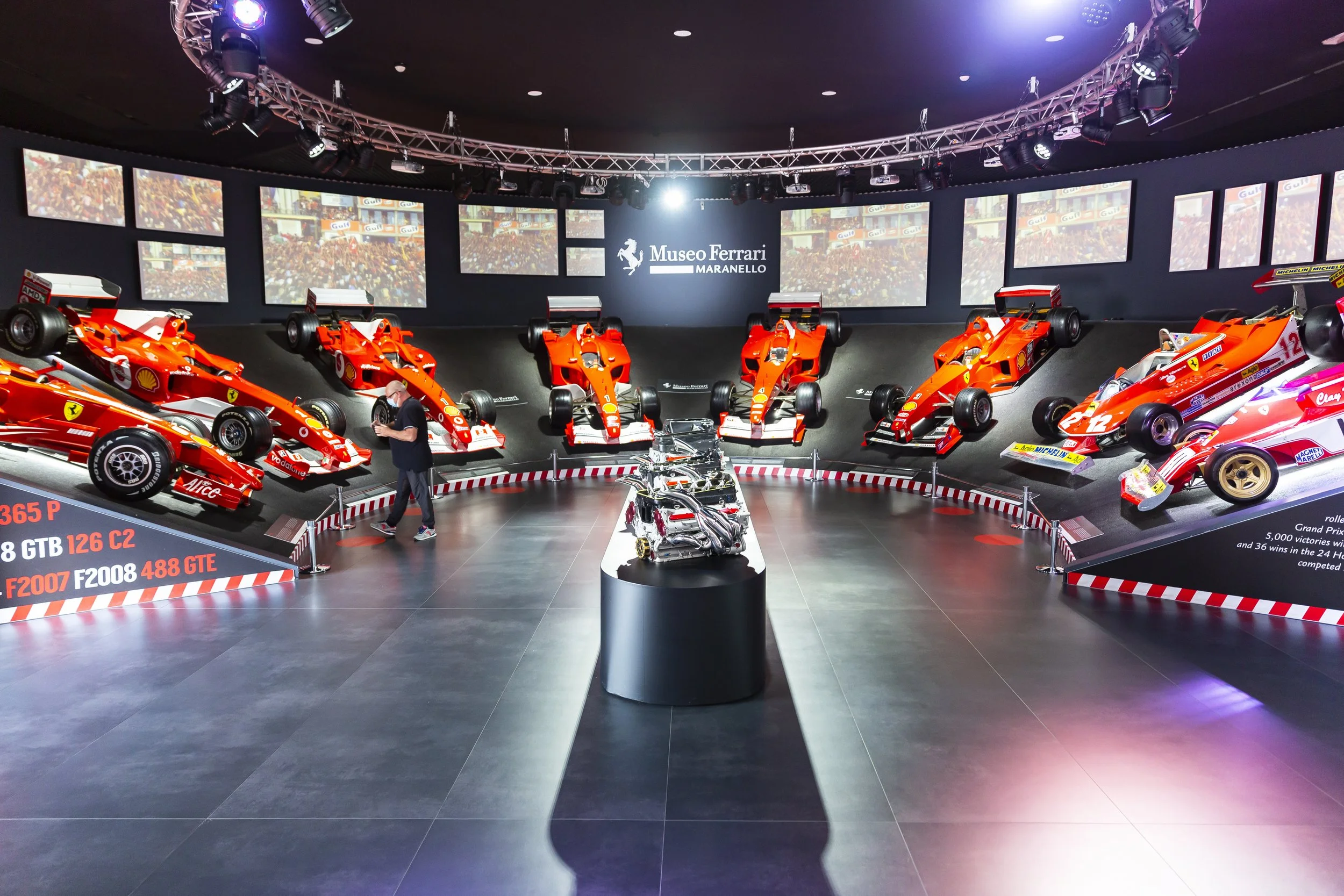 Exhibition of Formula 1 cars at the Ferrari Museum Maranello. Multiple red race cars on display, some mounted on inclined platforms, with a person viewing them. The museum has a dark interior, with a circular track around the cars, and large screens showing racing footage in the background.