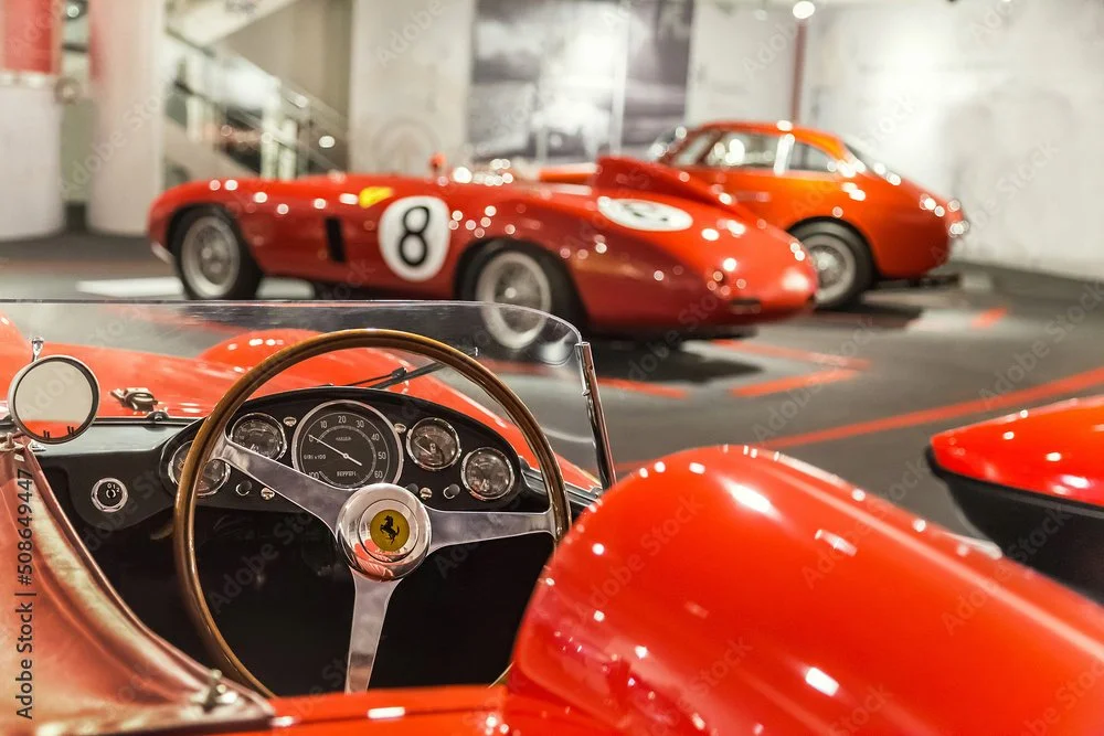 Interior view of a vintage Ferrari race car with a wooden steering wheel and classic dials, with two red racing cars displayed in the background.
