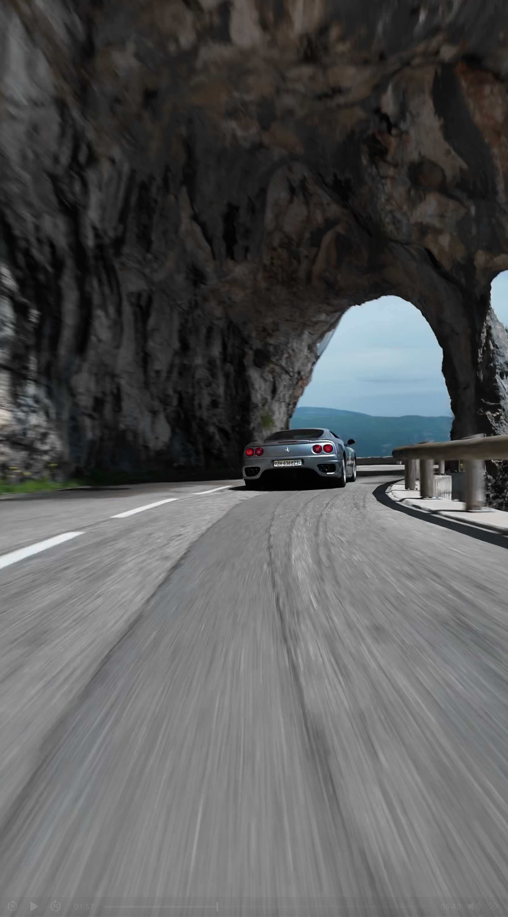 A silver sports car driving through a rocky mountain tunnel on a curved road with guardrails, distant mountains in the background.