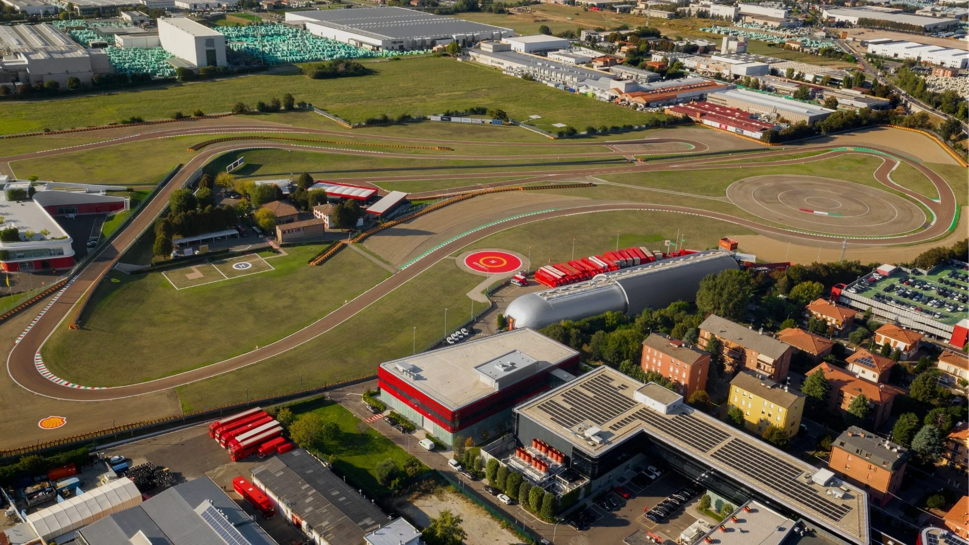 Aerial view of a racetrack with multiple curves, surrounding facilities, and nearby parking lots, with buildings and greenery in the background.