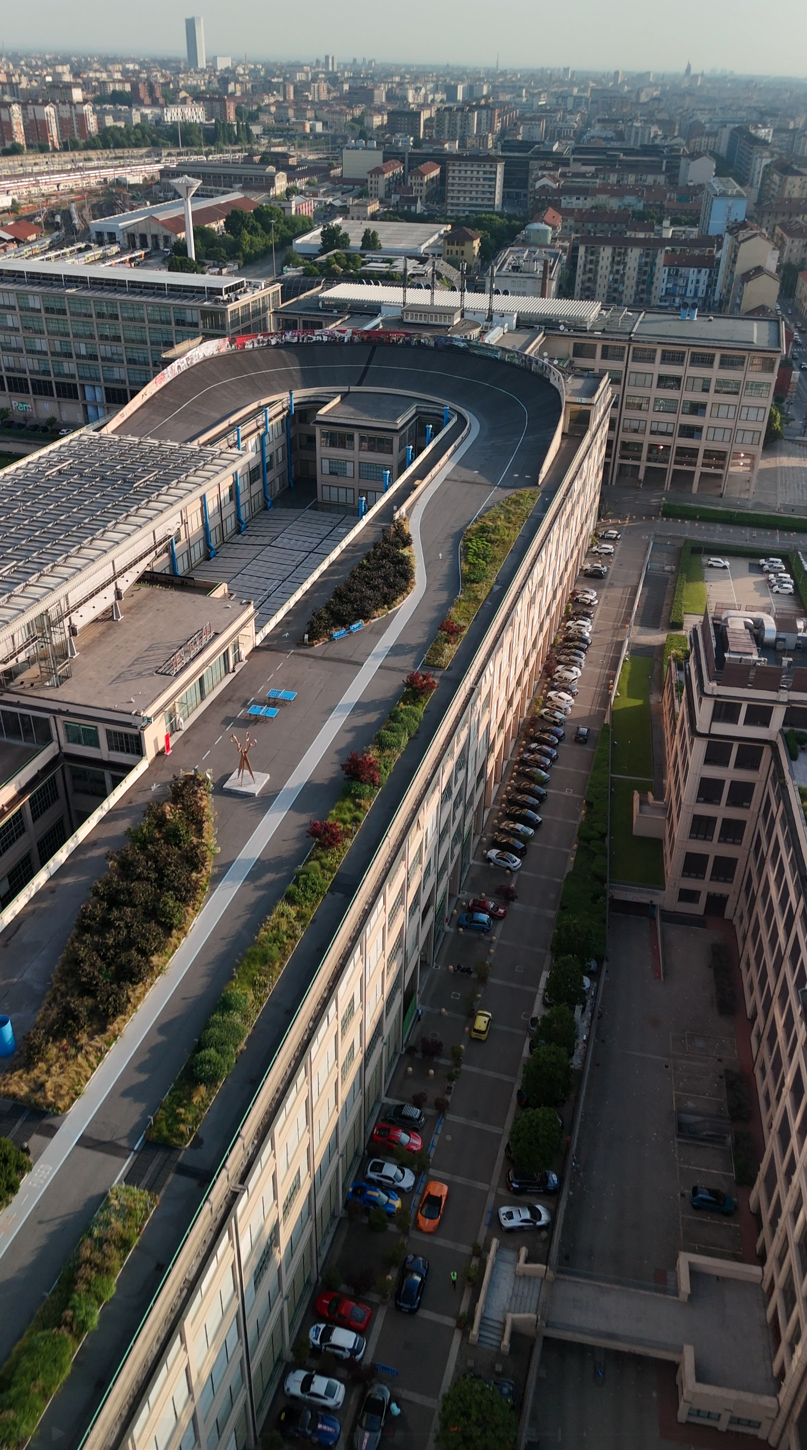 Aerial view of a cityscape showing a large building with a rooftop garden and parking lot, surrounded by other urban structures and streets.