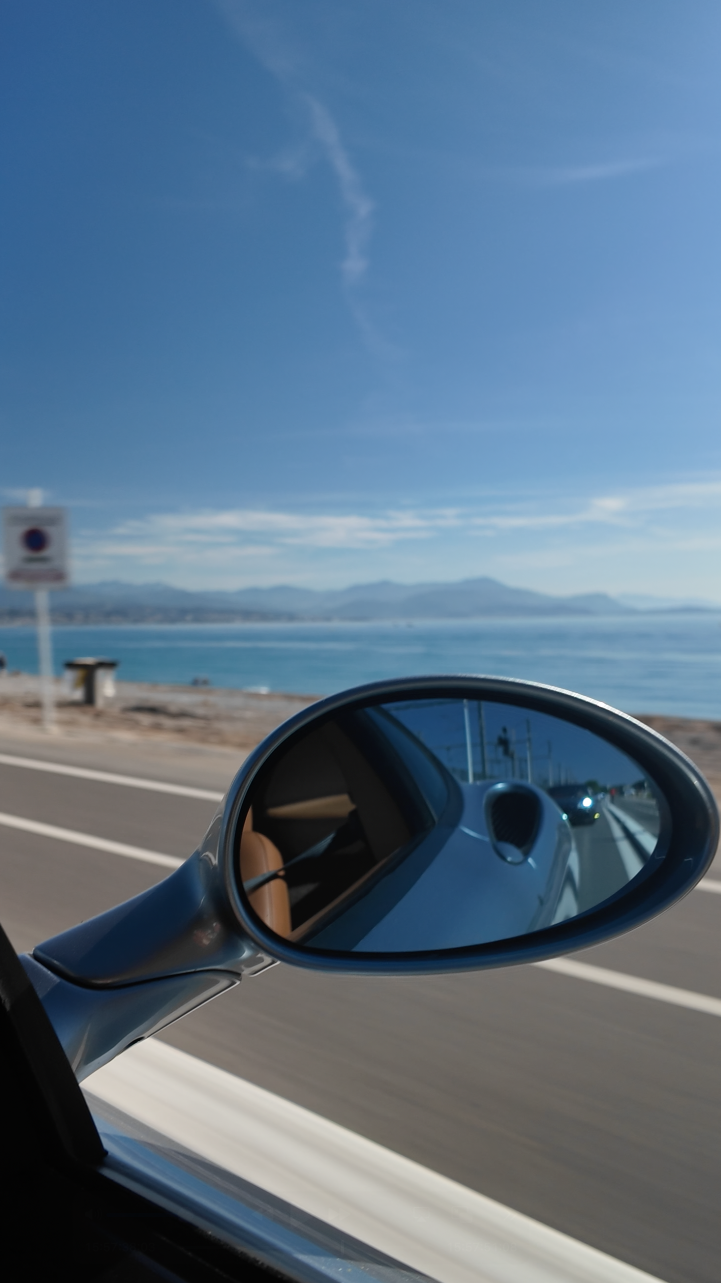 View of a seaside highway seen through a car's side mirror, with a blue sky, distant mountains, and water in the background.
