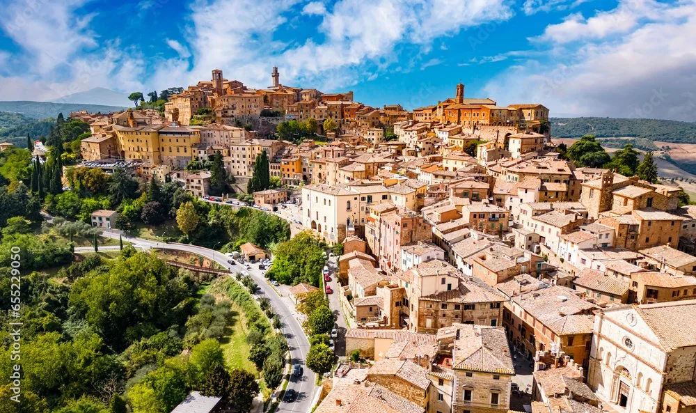A hillside town with tightly packed tan and brown buildings, winding streets, and green trees, under a partly cloudy sky.