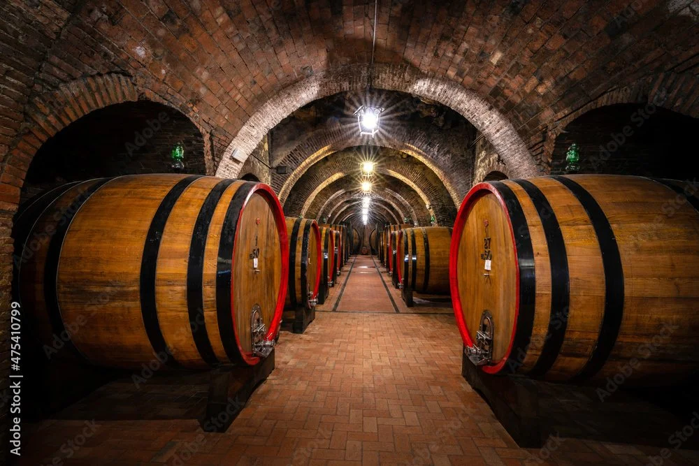View of a wine cellar with large wooden barrels arranged in two rows along a brick walkway, with brick arched ceilings and soft lighting.