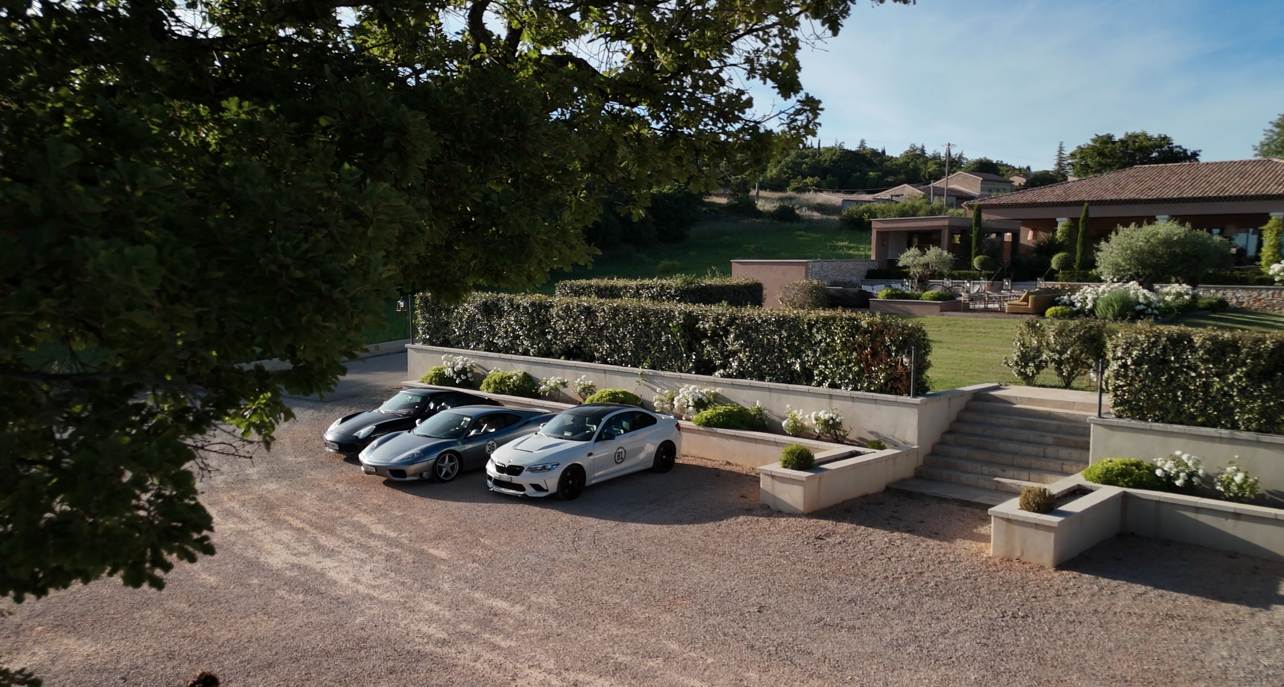 A view of a luxurious estate with a manicured lawn, staircases, and a variety of trees and shrubs. Four cars are parked in a gravel area in front of a white wall with decorative bushes. The background features a hillside with houses and lush greenery.