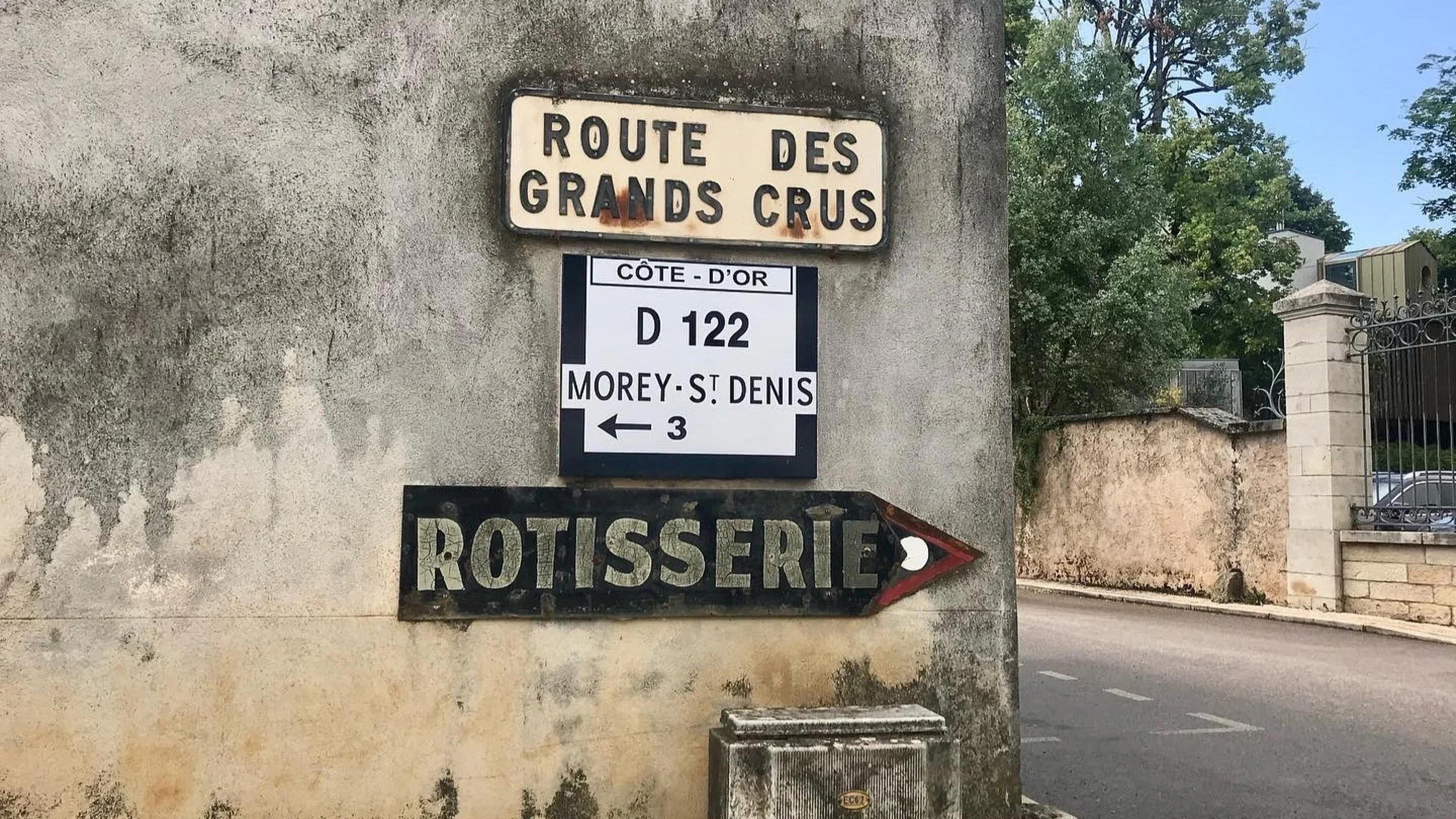 Wall with three signs: a white sign with black lettering reading "ROUTE DES GRANDS CRUS," a white parking sign with black text indicating "CÔTE-D'OR D 122 MOREY-ST DENIS 3" with an arrow pointing left, and a black sign with white lettering reading "ROTISSERIE" with a red arrow pointing to the right,T