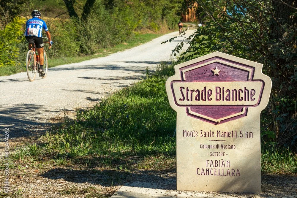 A gravel path along a wooded area with two cyclists riding on it. A sign reads "Strade Bianche, Monte Sante Marie 1.5 km, Comune di Asciano, Settore, Fabián Cancellara."