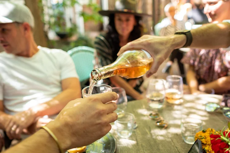 A group of people sitting around a table at a gathering, with one person pouring what appears to be wine or a similar beverage into a glass. The setting appears to be cozy and outdoors, with natural light and greenery in the background.