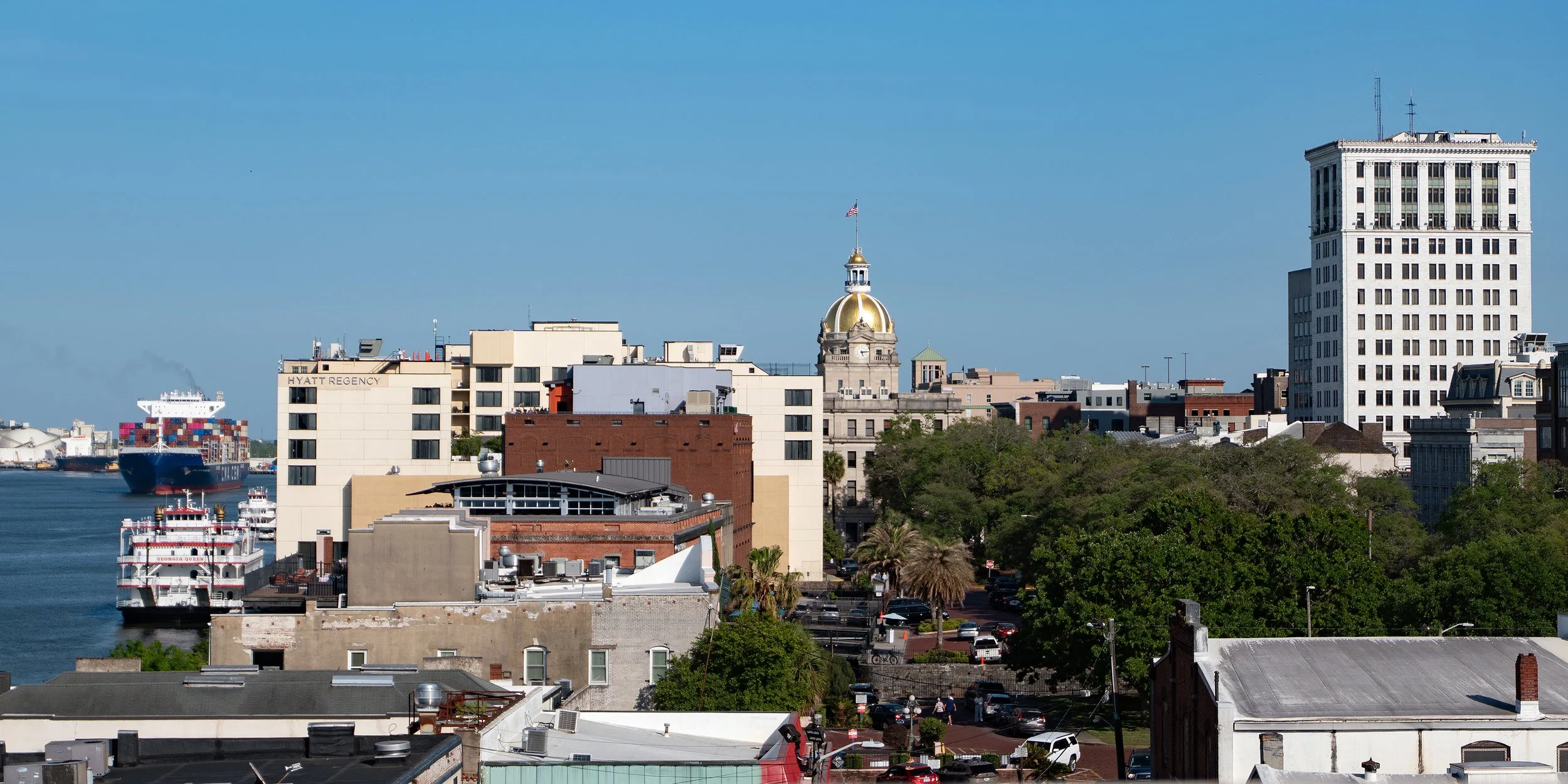 View of the Savannah skyline featuring the Georgia State Capitol dome, the Georgia Queen riverboat, and a cargo ship on the Savannah River from The Lost Square rooftop.