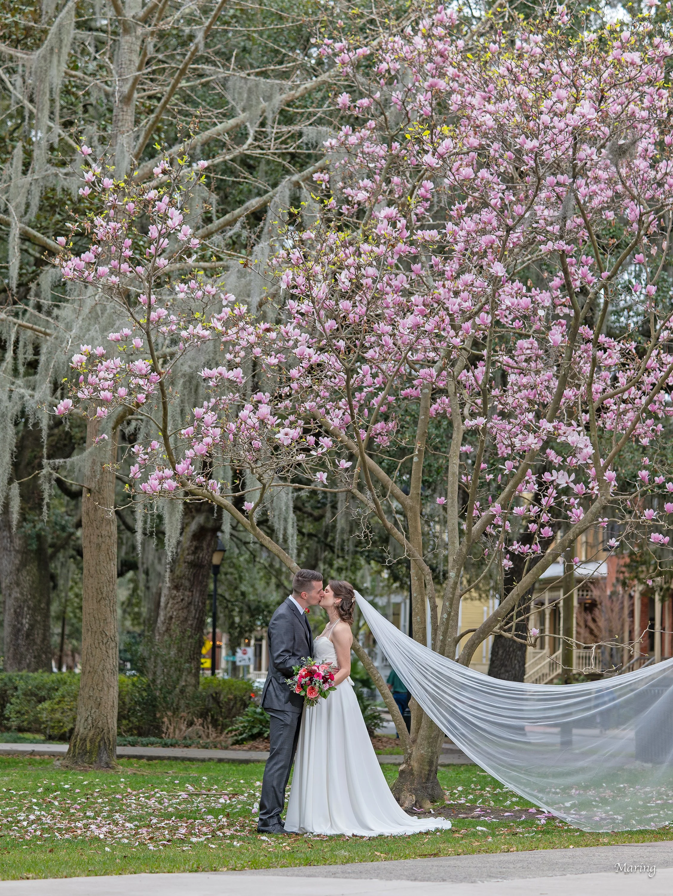 Bride and groom pose for a spring wedding portrait in Forsyth Park surrounded by blooming floral trees during a romantic Savannah wedding day.