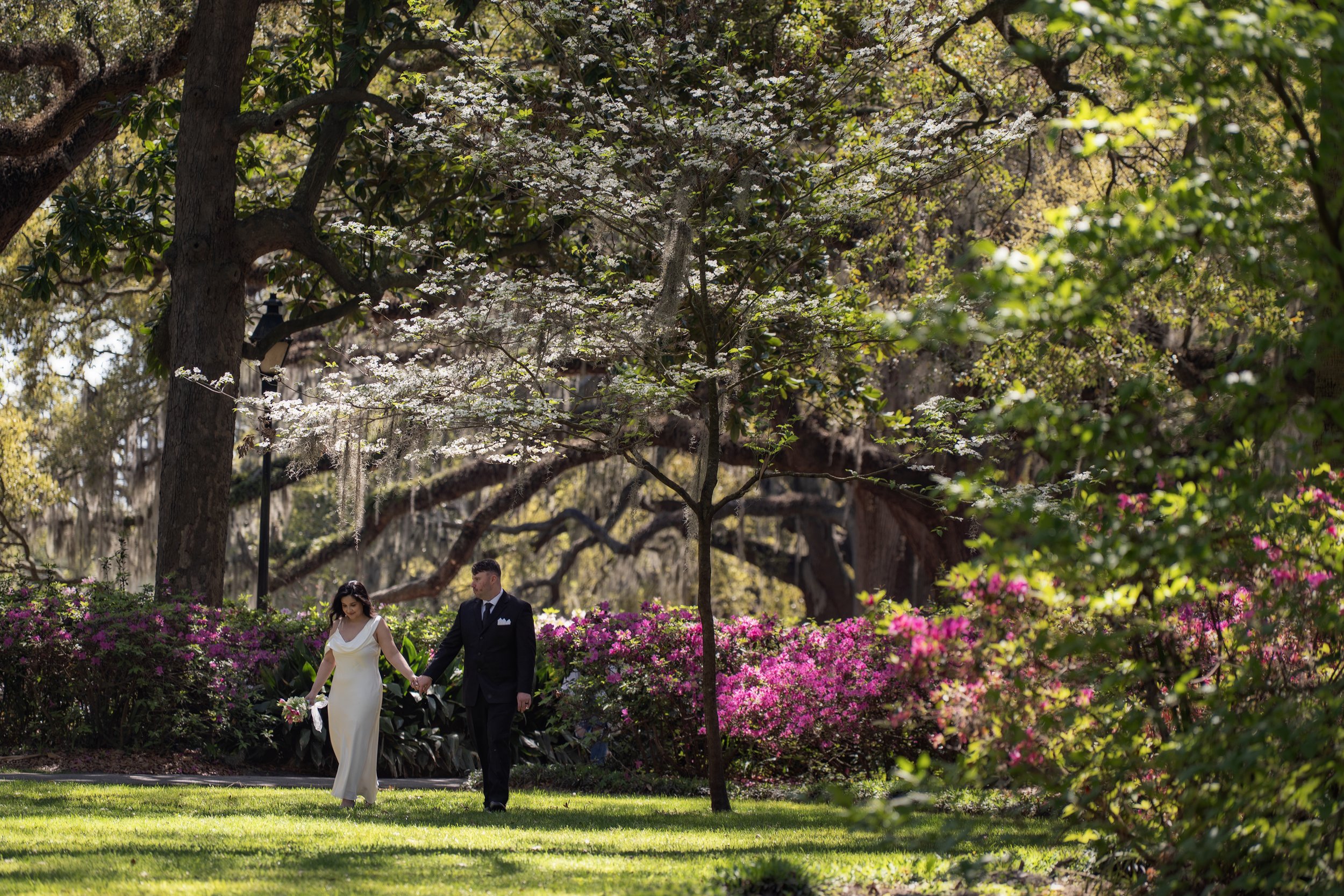 Bride and groom walking hand in hand through Forsyth Park surrounded by blooming trees and azaleas in Savannah Georgia.