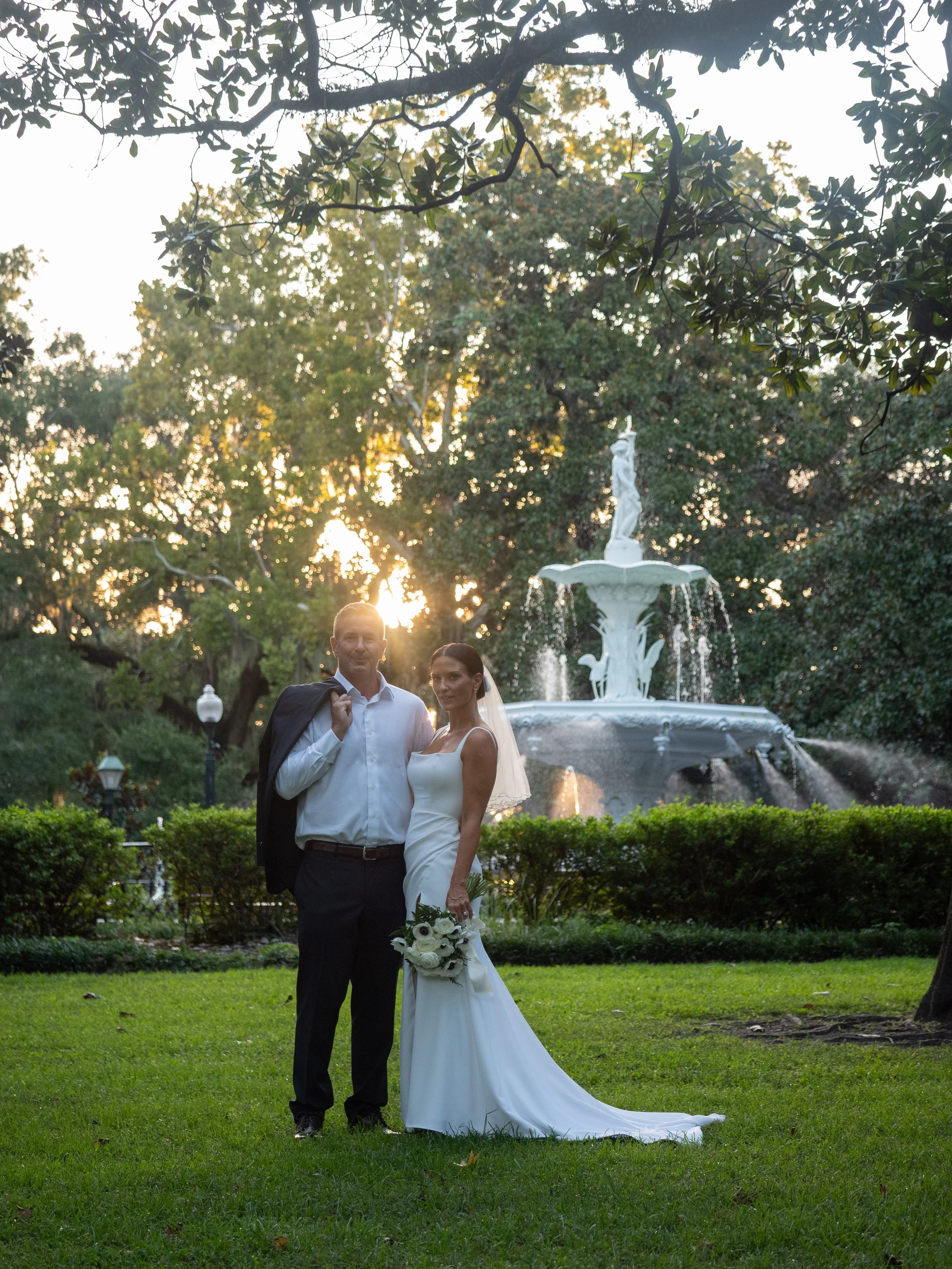 Bride and groom standing in front of Forsyth Park Fountain with sunlight filtering through the trees