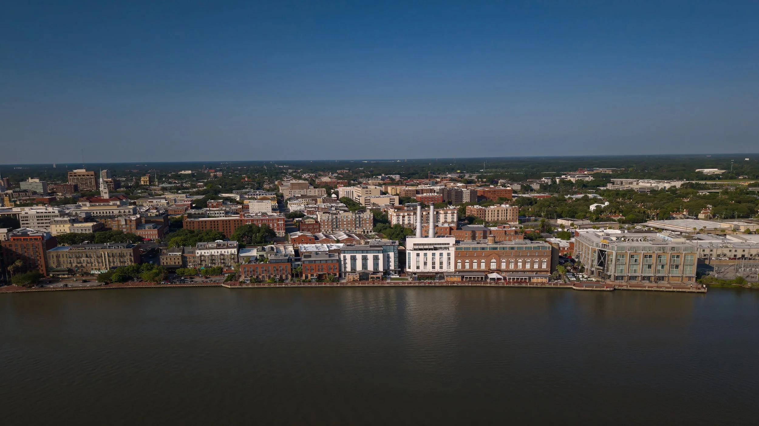 Aerial view of the Savannah Ga skyline along a river under a clear blue sky, featuring a mix of modern and historic buildings.