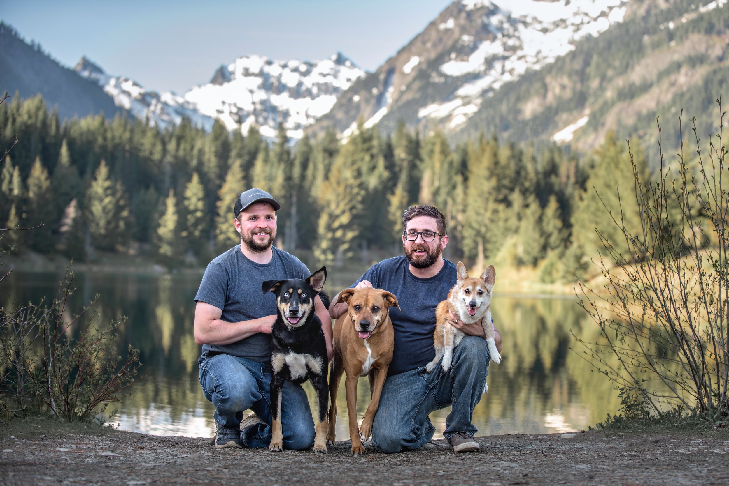 A gay couple kneeling with their three dogs in front of a stunning lake and mountain view