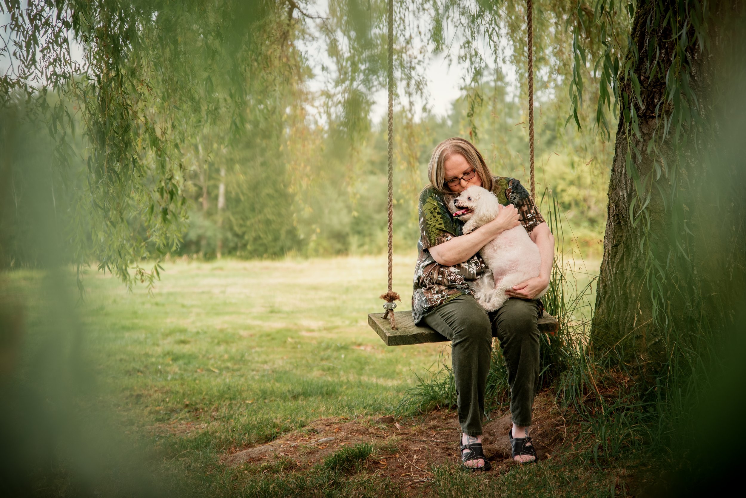 Senior dog mom cuddling senior maltese while sitting on a wooden swing hung from a willow tree in Tacoma