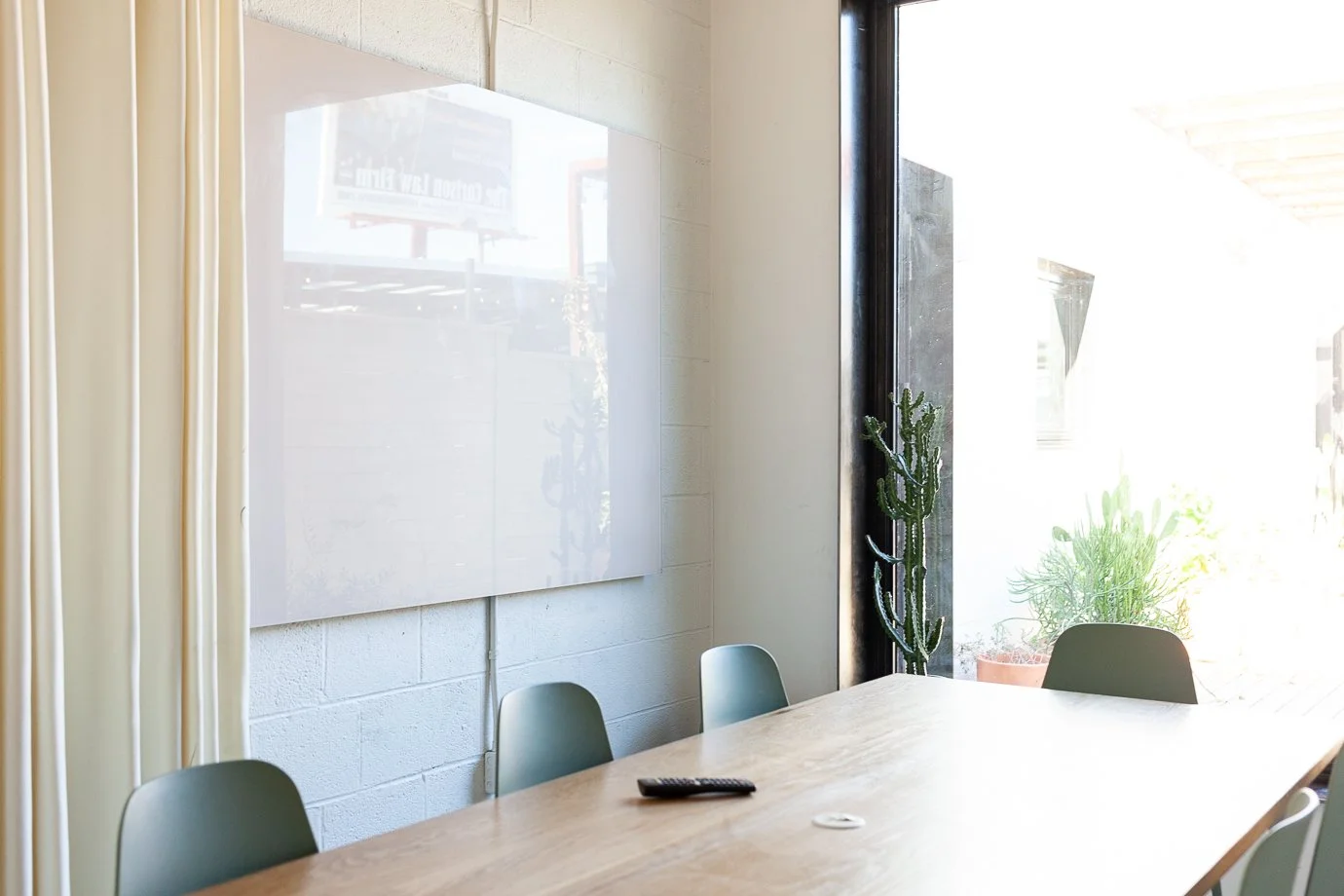 Empty conference room with a wooden table, green chairs, a remote control, and a whiteboard, next to large windows with potted plants outside.