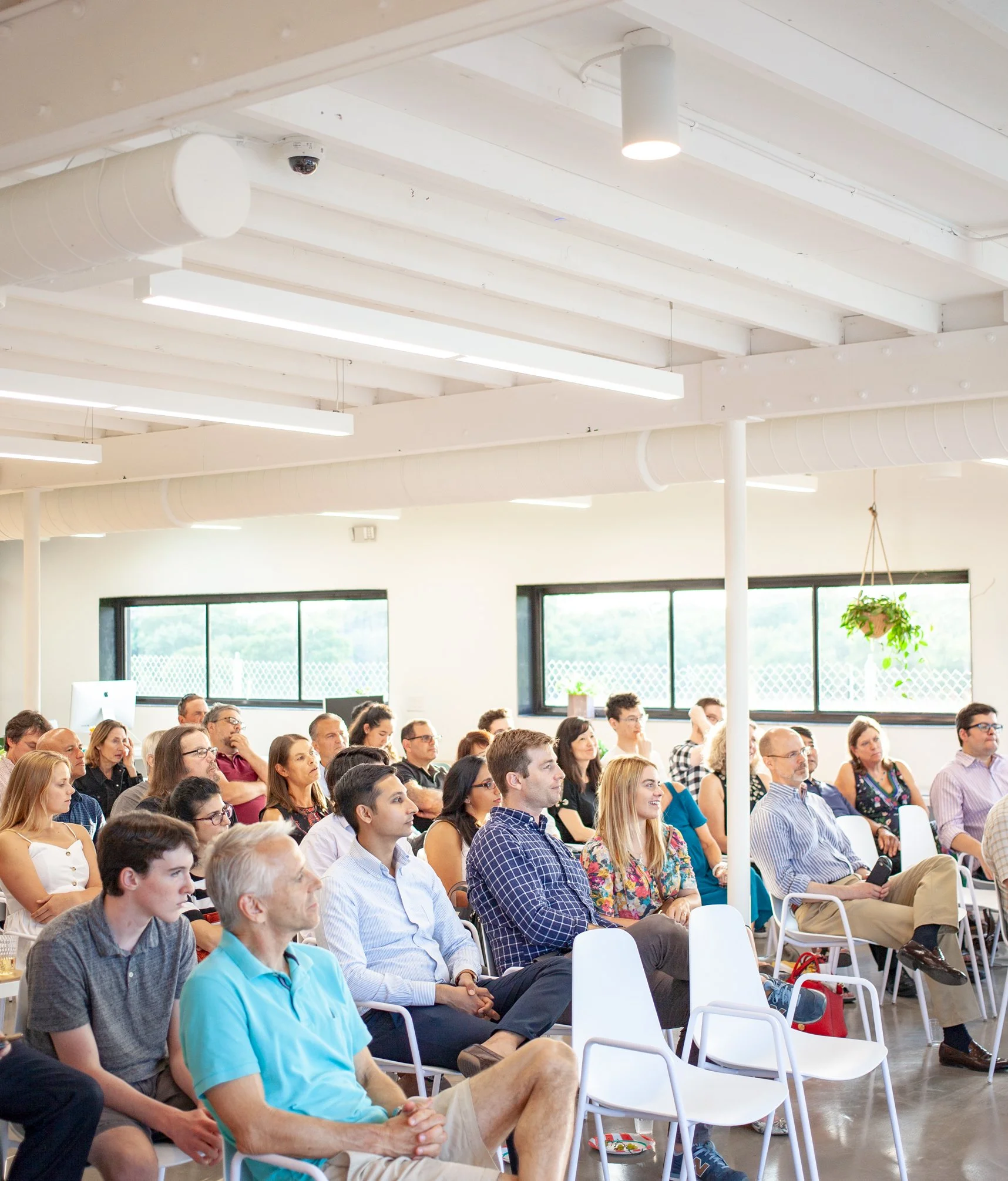 A group of people attending a presentation or conference in a modern, well-lit room with large windows and hanging plants.