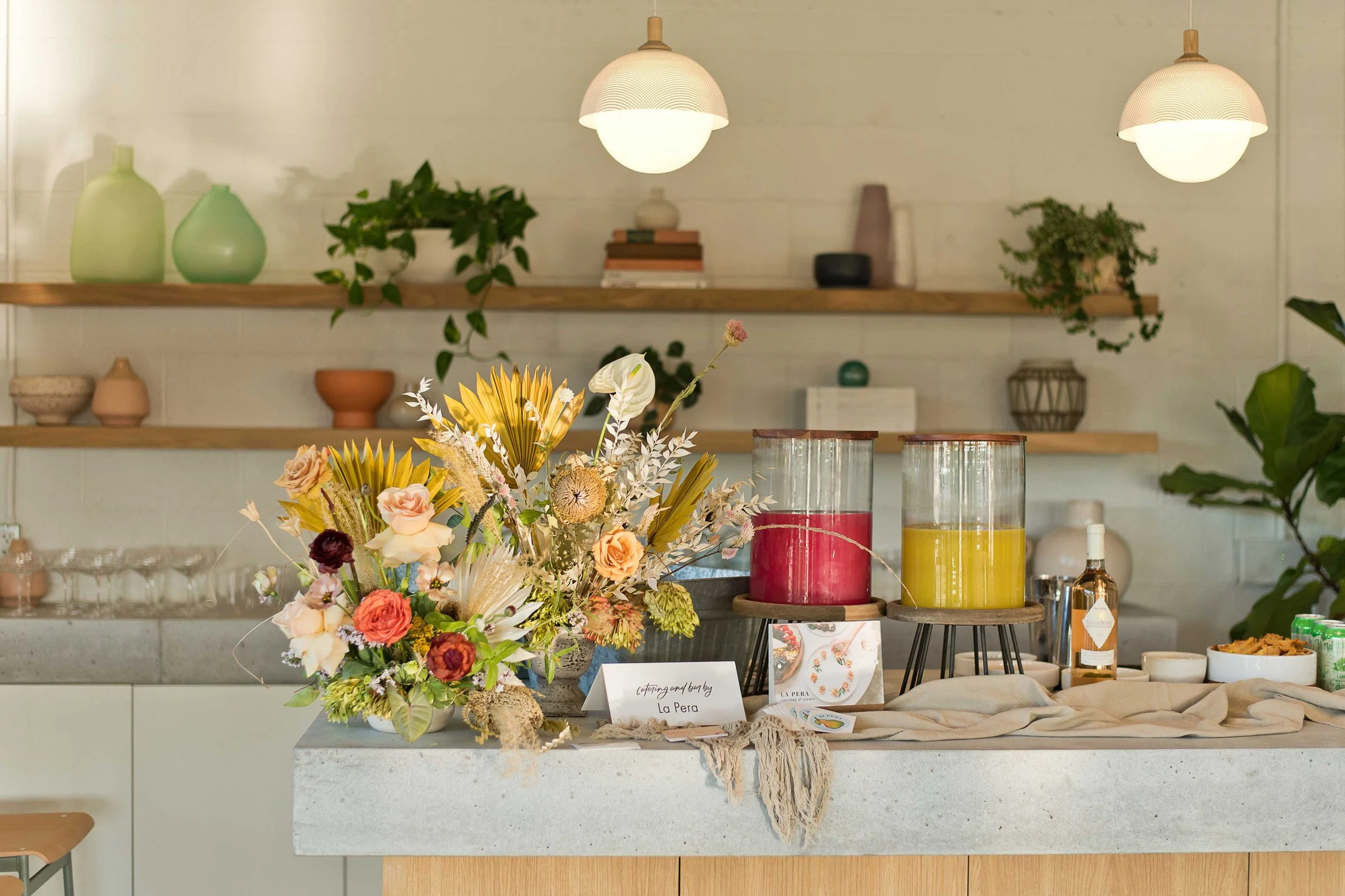 Decorative display on a kitchen island with a large colorful flower arrangement, two cylindrical glass beverage dispensers with red and yellow drinks, and various small items. In the background are wooden shelves with vases, plants, and decorative objects, illuminated by white pendant lights.