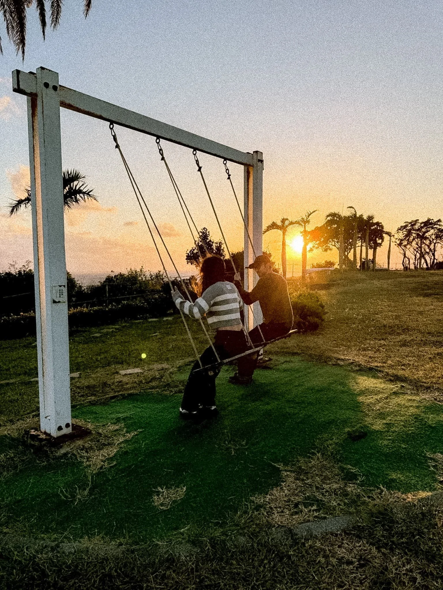 Person sitting on a swing at golden hour at SkyeBay Club resort in Kenting Taiwan with palm trees and sunset sky