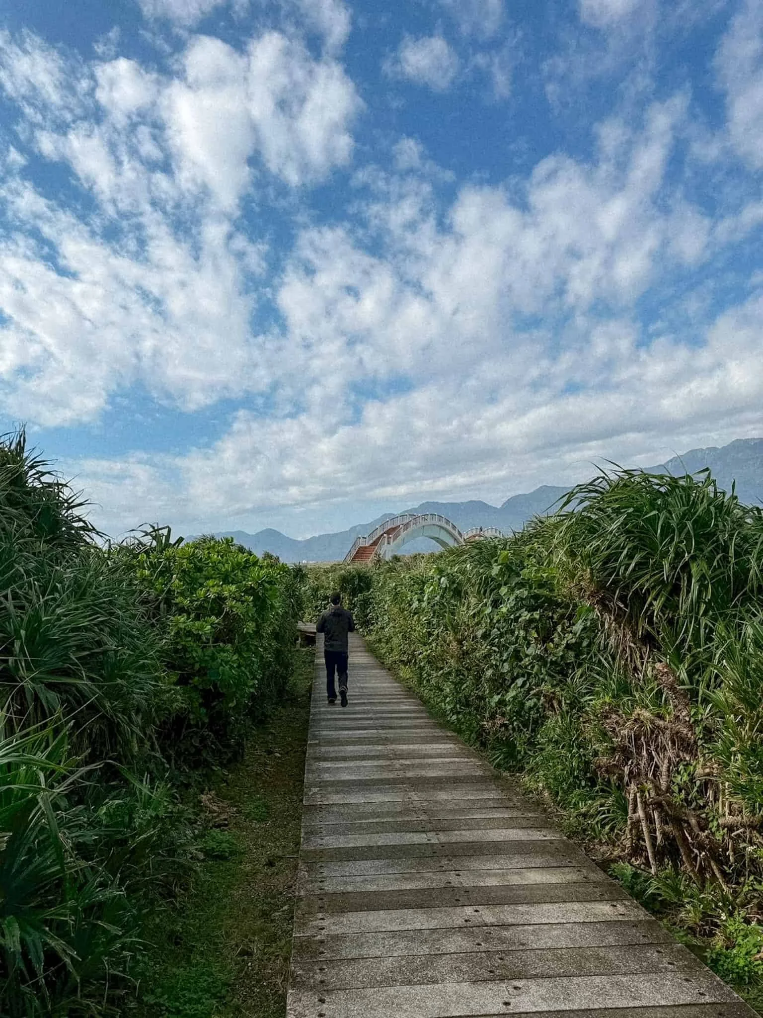 Wooden boardwalk trail on Sanxiantai island winding through coastal shrubs toward the bridge, Taitung