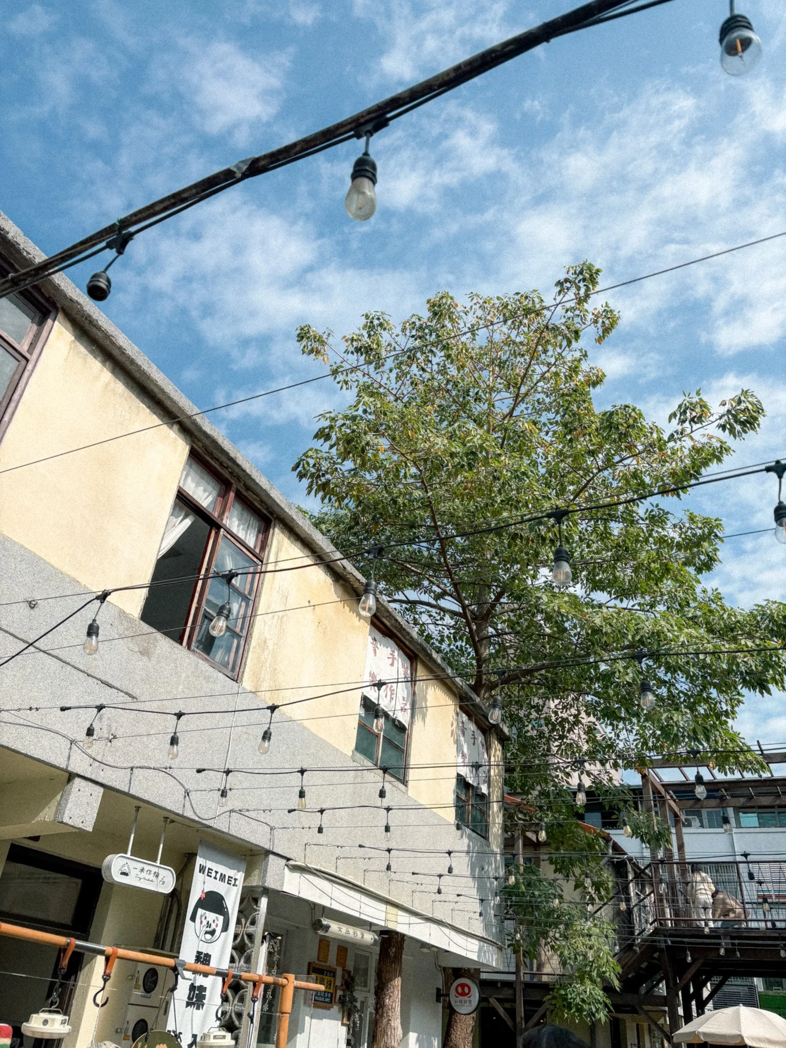 String lights and a leafy tree above restored old buildings and small shops in Shengji New Village, Taichung