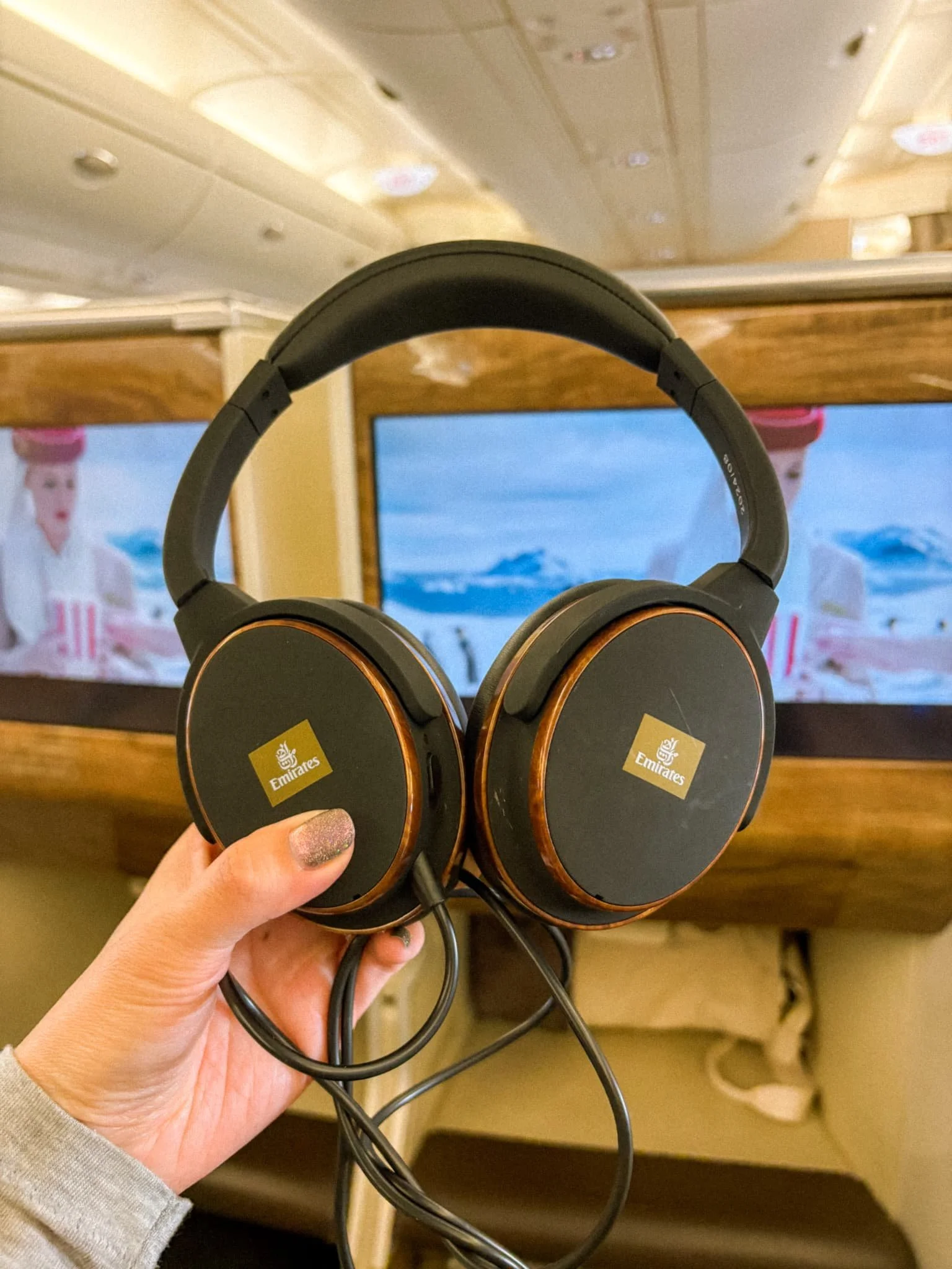 Emirates Business Class noise canceling headphones held up in front of the in-flight entertainment screen on the A380