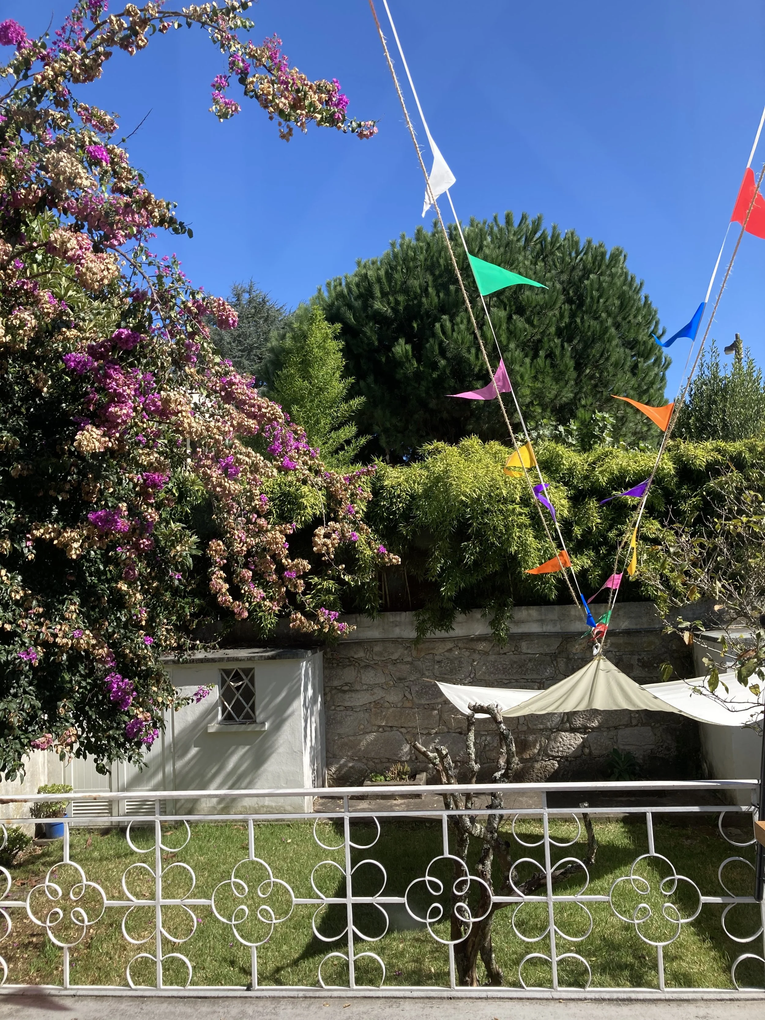 Jardim com árvores floridas, árvore em formato de broto e uma estrutura de guarda-chuva com bandeirinhas coloridas suspensas, céu azul claro no fundo.