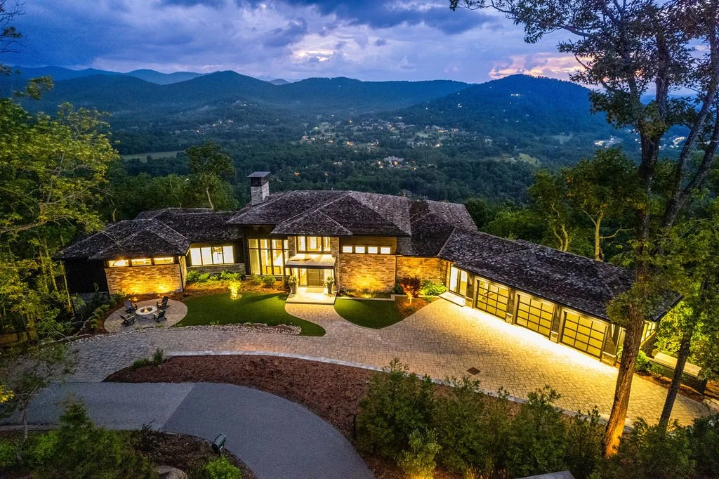 Aerial twilight view of a luxury mountain home with stone siding, illuminated windows and panoramic Blue Ridge views near Asheville, NC.