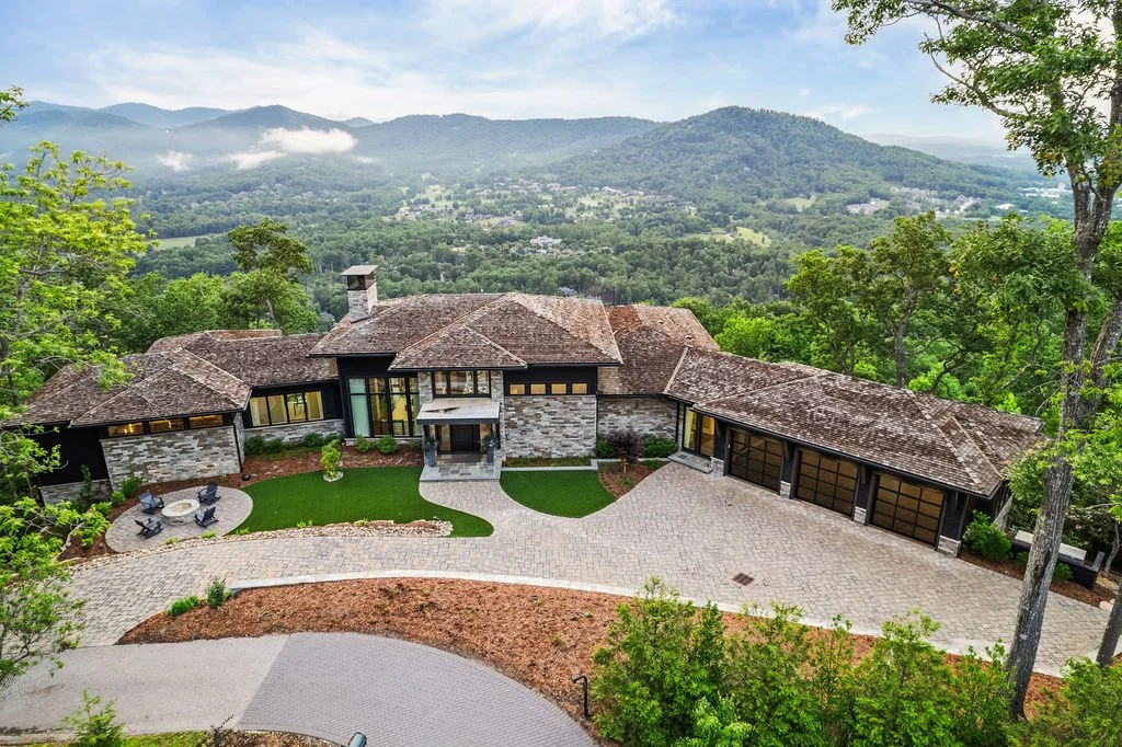 Aerial view of a luxury custom home with stone and dark siding, landscaped grounds, a fire pit and panoramic mountain views in the Carolinas.