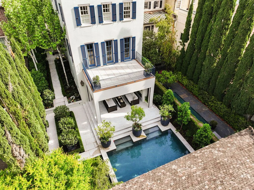 Aerial photo looking down into lush backyard with a three-story white house with a balcony, patio and pool surrounded by trees.