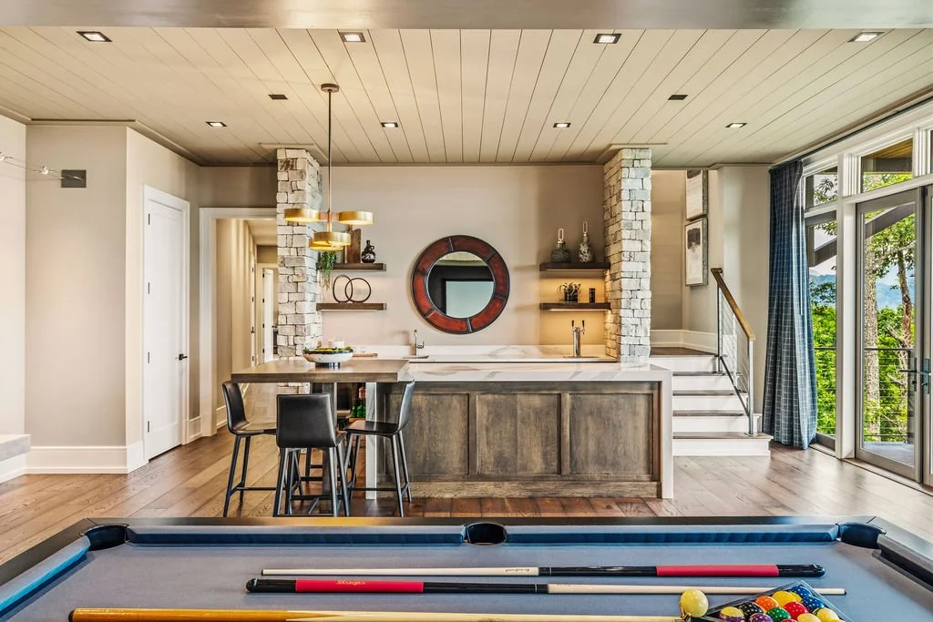 Home bar with seating at one end, a hall leading away, shelves on the wall, stairs leading up on the right side, large windows on the edge of the photo and a pool table in the bottom foreground.