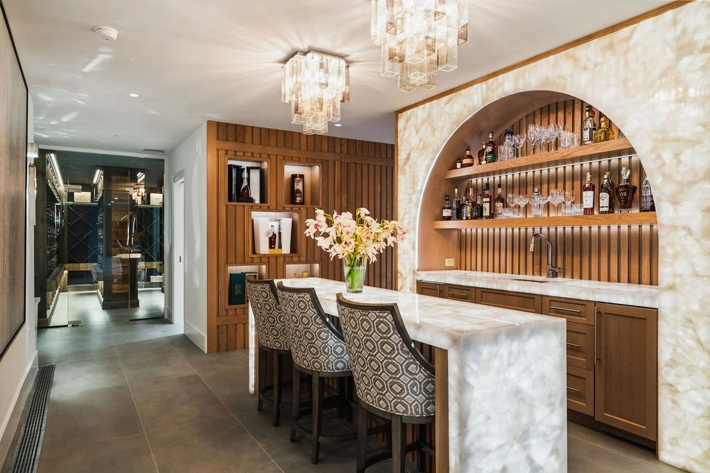 Luxury home bar with glowing marble countertops and backsplash, three seats, liquor bottles and a door leading to a wine cellar.