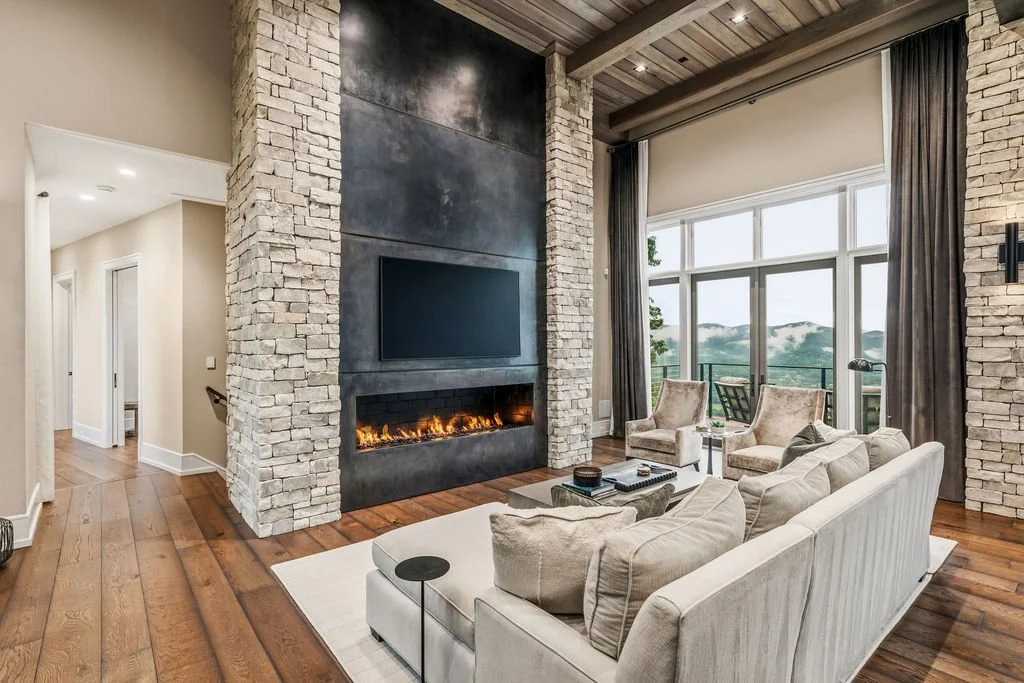 Modern living room with high ceilings, floor-to-ceiling windows, a couch facing a fake fireplace with a tv above it and a hall on the left side leading away.