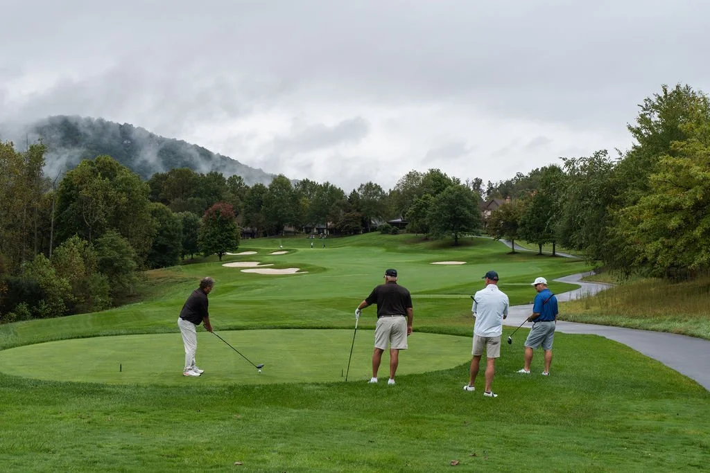 Group of three men watching another man hit a golf ball on a green fairway with sand traps on a foggy day
