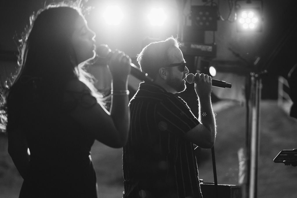 Black-and-white image of a woman and a man in sunglasses singing into microphones with stage lights behind them.