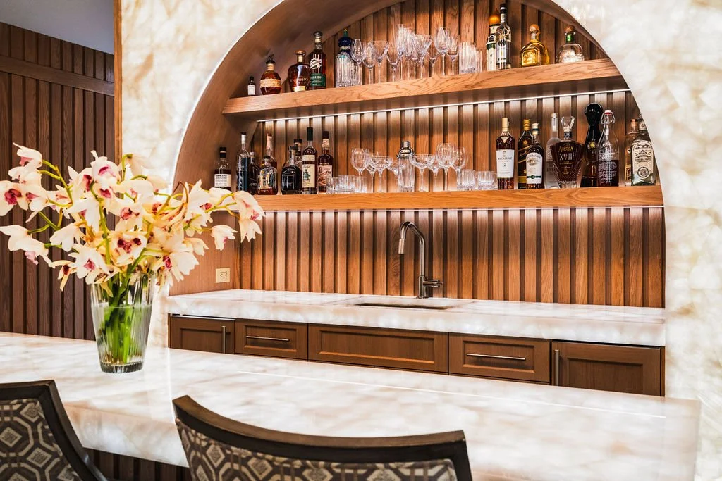 Custom home bar with arched wood shelving, integrated lighting, translucent stone surfaces and a marble-topped island.