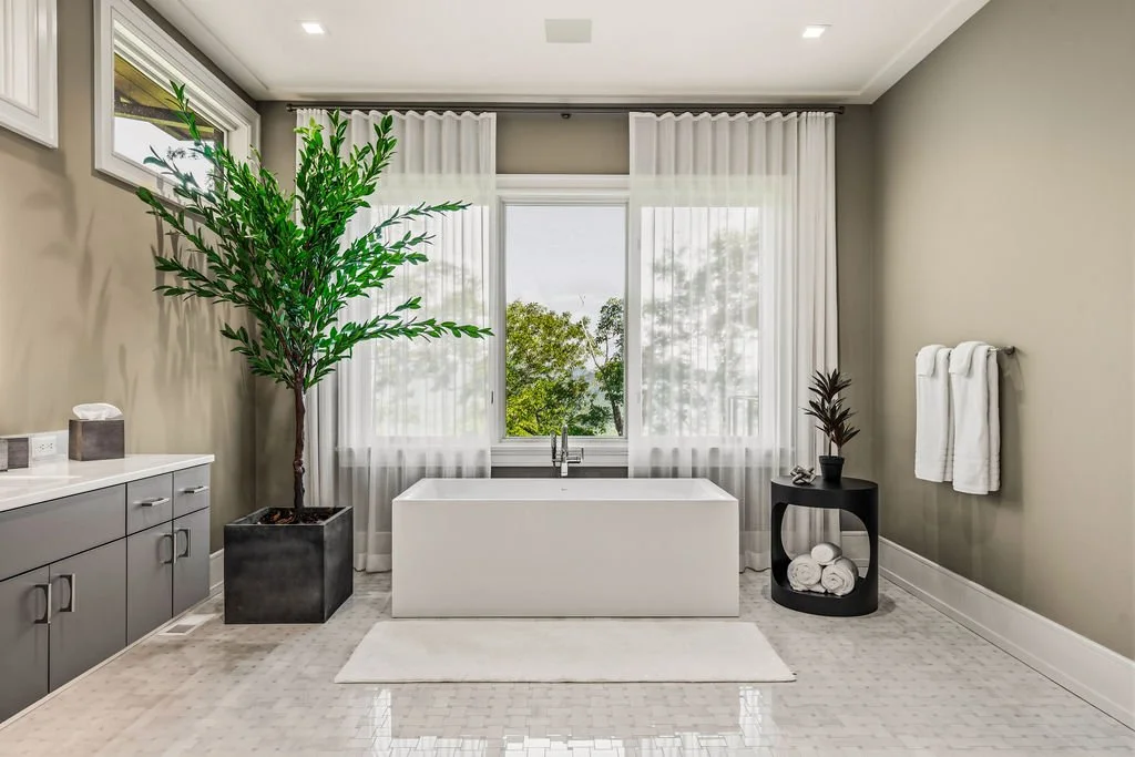 Bathroom with white freestanding tub, large windows with transparent curtains, potted tree, side table, towels and countertop.
