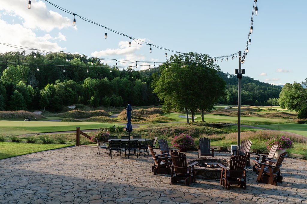 Outdoor patio with fire pit, table, chairs and string lights with a lush green golf course behind it and blue skies with a few clouds.