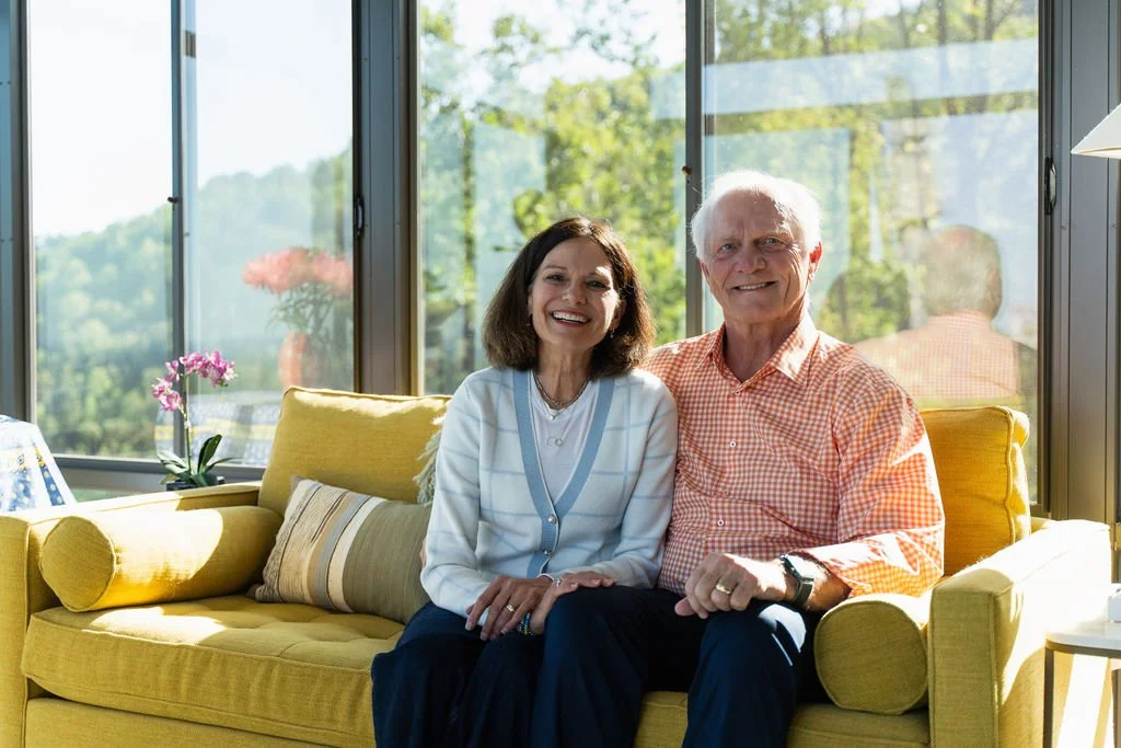Lifestyle portrait of a smiling elderly couple seated on a yellow sofa in a bright modern home with large windows and wooded views.