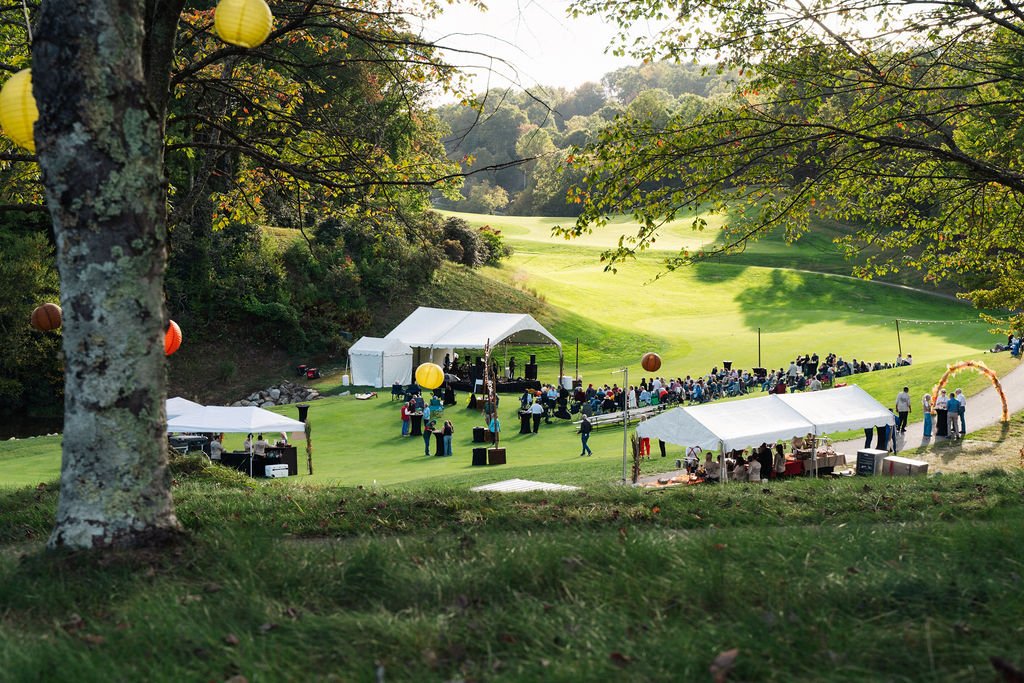White festival tents setup in the valley of a golf course surrounded by trees and the top of a hill and tree in the foreground.