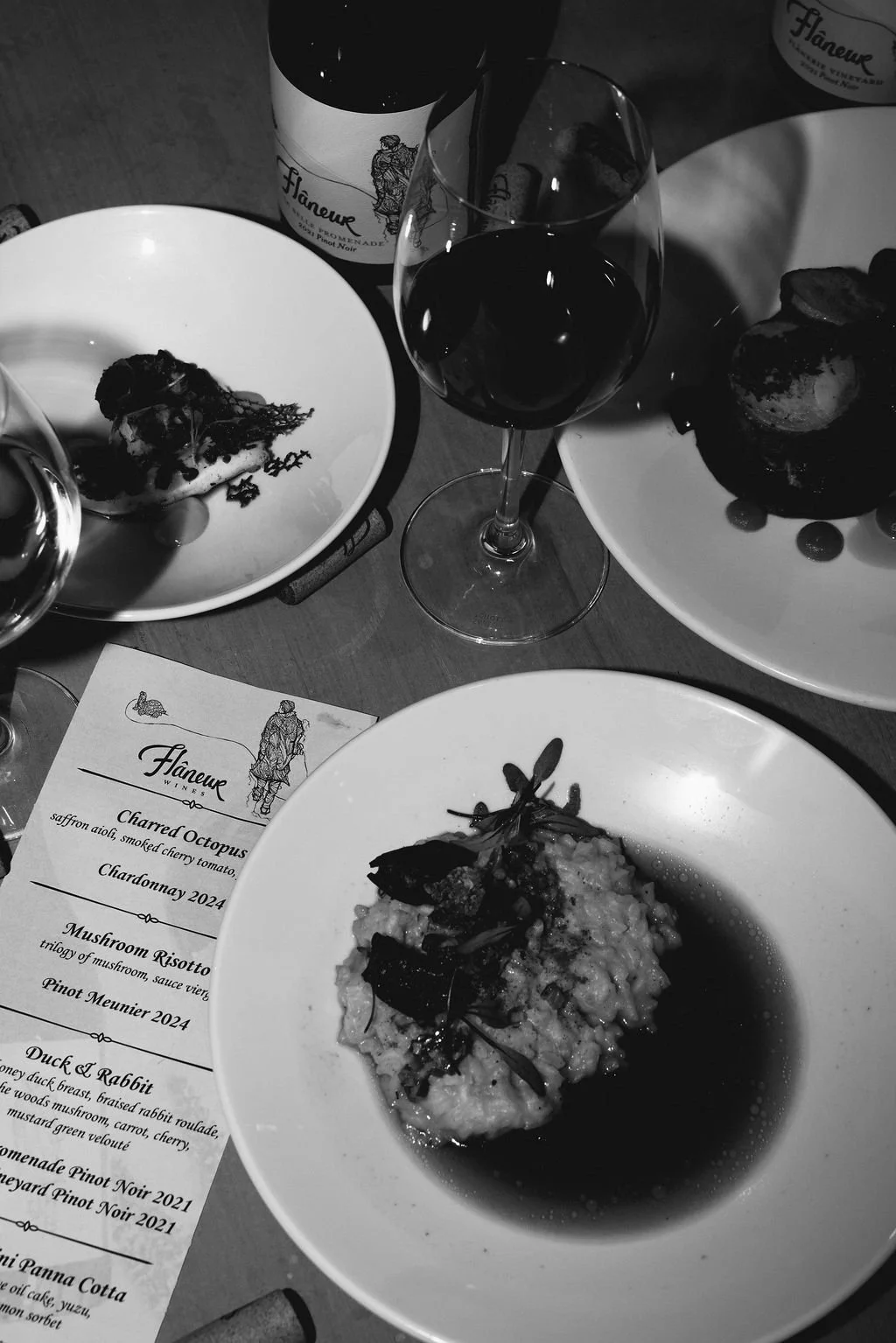 Black-and-white image of fine dining table with three plates of food, a menu, wine bottle and two glasses.