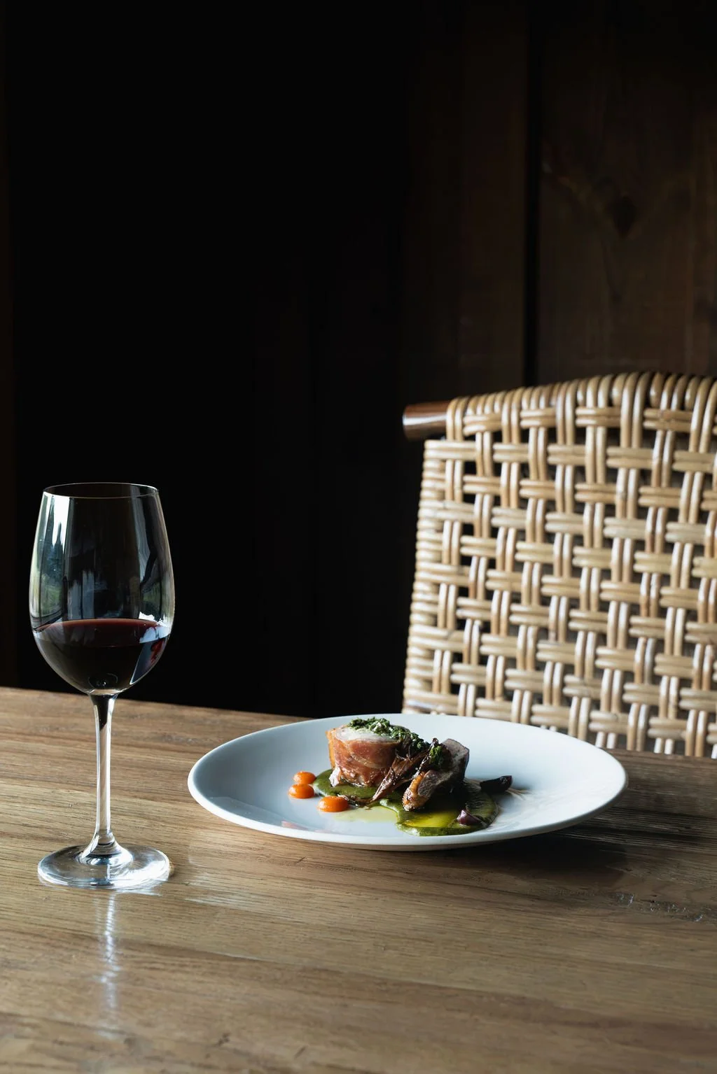 Professional restaurant food photography of an elegant plated entrée beside a glass of red wine in a warm, modern dining space.