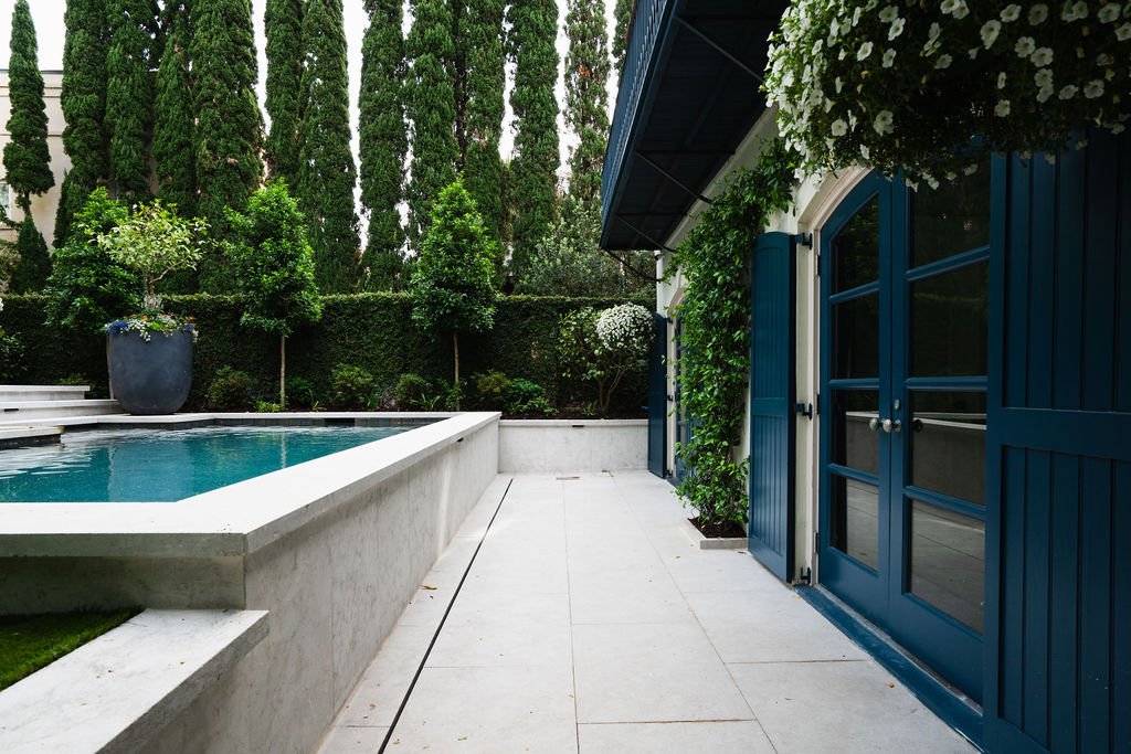 Charleston courtyard pool beside a white stucco home with deep blue shutters, arched doors, formal hedges and tall cypress trees.