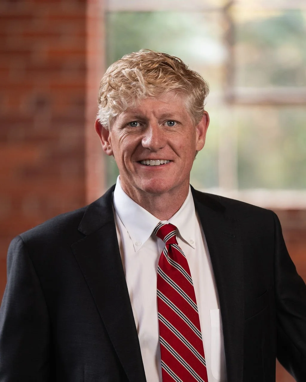 Professional business headshot of a man in a dark suit and red striped tie with a blurred brick wall and window in the background.