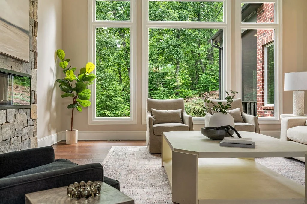 Luxury living room with a stone fireplace, neutral furnishings, hardwood floors and tall windows overlooking a wooded landscape.