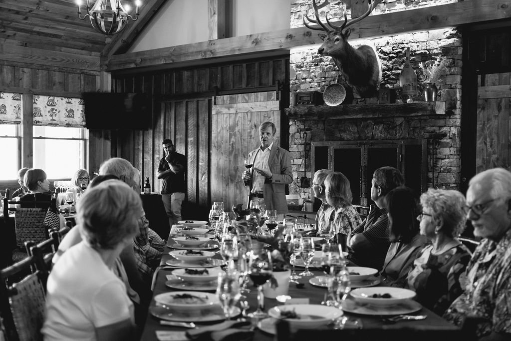 Black-and-white image of a wine dinner with a long table and a man giving a toast at the end in a room with wood beams, stone fireplace and a deer mount.